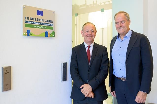 Patrick Child, stellvertretender Generaldirektor der Generaldirektion Umwelt bei der Europäischen Union (EU) (l.) und Oberbürgermeister Eckart Würzner stehen neben dem EU-Mission-Label „Klimaneutrale und intelligente Stadt“ im Heidelberger Rathaus. (Foto: Philipp Rothe)