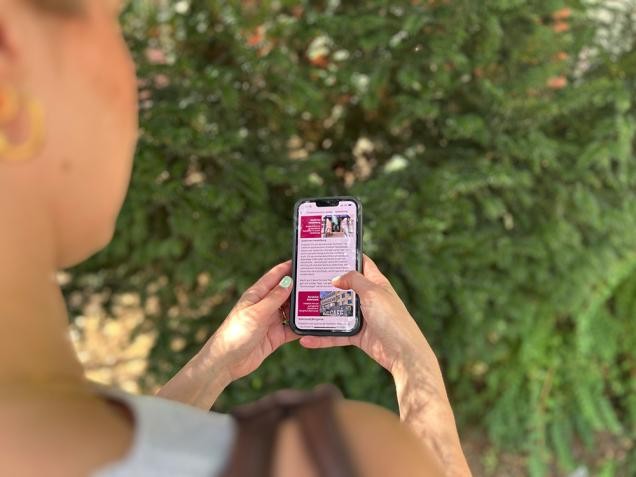 Blick über die Schulter einer jungen Frau, sie bedient ein Smartphone mit geöffneter MeinHeidelbergApp, im Hintergrund ist verschwommen ein Busch zu sehen.