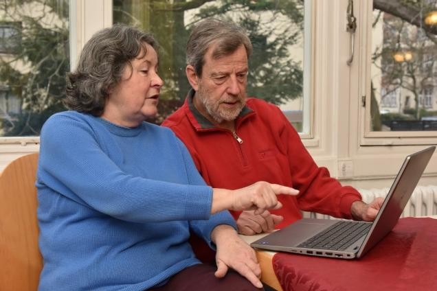 Zwei ältere Menschen (eine Frau in blauem Pulli und ein mann in rotem Pulli) sitzen mit einem Laptop an einem Tisch. Die Frau zeigt mit ihrem Finger auf den Bildschirm, beide schauen in die Richtung des Bildschirmes.