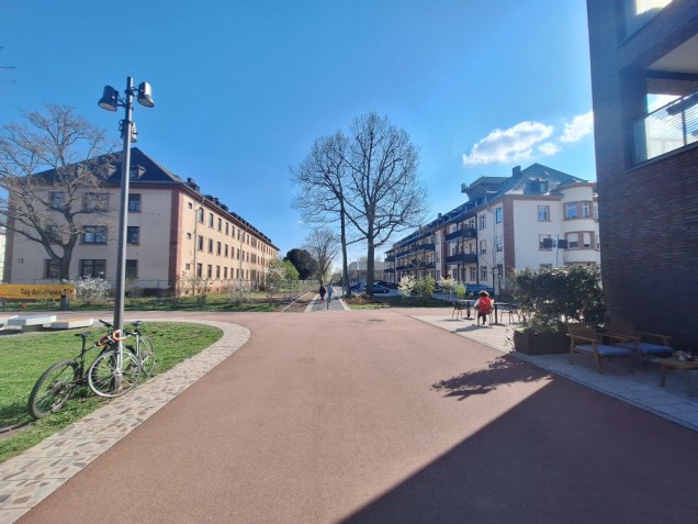 Das verkaufte Gebäude (links) liegt zentral in der Nähe des Paradeplatzes und am Fußweg der Adelheid-Steinmann-Straße. Es wird künftig die Heimat des gemeinschaftlichen Wohnprojektes paraSol sein. (Foto: Entwicklungsgesellschaft Campbell Barracks mbH) Zu sehen ist ein breiter Weg der nach links abbiegt. Weiter geradeaus liegen zu beiden Seiten 2 große Gebäude.