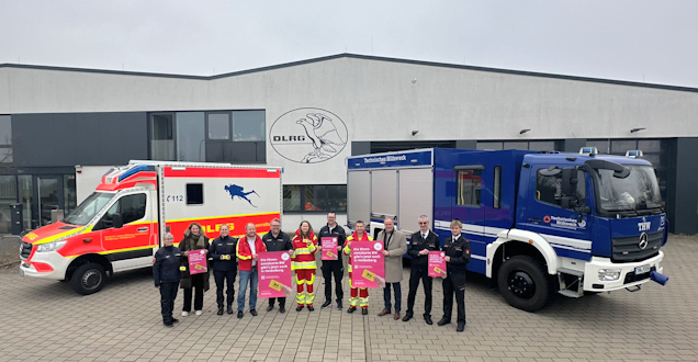 Gruppenfoto von Angehörigen von Feuerwehr, DLRG und THW mit OB Würzner vor Einsatzfahrzeugen. Die Teilnehmer halten Infoplakate zu Ehrenamtskarte in die Kamera.