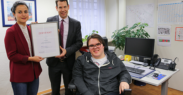 Eine Frau und ein Mann halten eine Urkunde hoch. Neben ihnen ist ein junger Mann im Rollstuhl. (Foto: Stadt Heidelberg)
